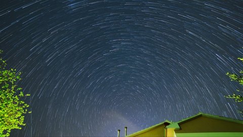 The stars move around the North Star. Trees and the roof of a house in the foreground. Time lapse of star trails in the night sky in Italy.