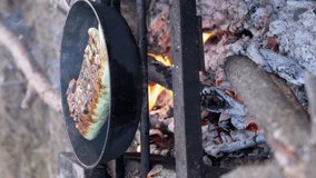 VERTICAL VIDEO: Cooking Meat Shawarma Wrapped in Pita Bread on Open, Burning Bonfire, in Pan. Male hands turn over the burnt, crispy lavash in blazing, smoking flame. Concept of food, hiking.
