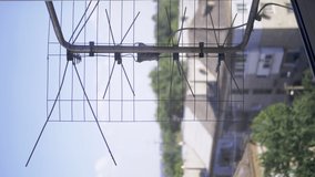 VERTICAL VIDEO: Old, Rusty TV Antenna is Attached to a Bracket Outside Window. A metal outdoor antenna is behind balcony a blurry background of sky, trees, clouds. Signal reception. Building facade.