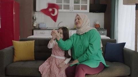 Istanbul,Turkey - 04.22.2022: A Turkish family is smiling and waving the Turkish flag in the living room. Mother and daughter in hijab celebrate their national holiday by waving the Turkish flag.