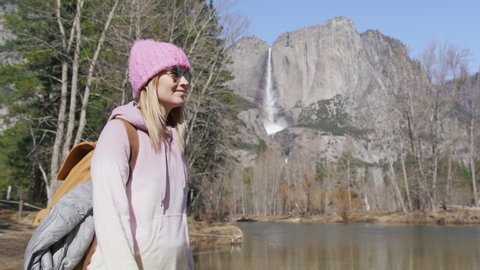 Slow motion relaxed woman enjoying Yosemite valley with waterfall and Merced river view. Active female with backpack on nature hike. Female traveler walking along dry trees forest, leisure bio-tourism