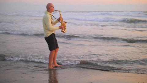 In summer, young bald man saxophonist plays golden alt saxophone at sunset, on seashore, beautiful waves with white foam, against sea, in yellow sweater, black shorts. Slow motion video. Romance.