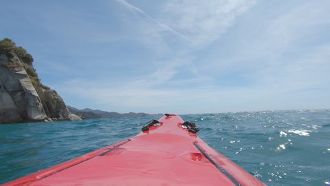 Red sea kayak splashing water towards the camera while crossing over waves in deep blue sea on a sunny day close to the coastline of Abel Tasman National Park 