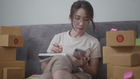 Video of a young beautiful woman sitting on the sofa and checking the costumer’s details from mobile phone and writing to the note book with a lot of parcel boxes around her. Soft mood and soft focus.