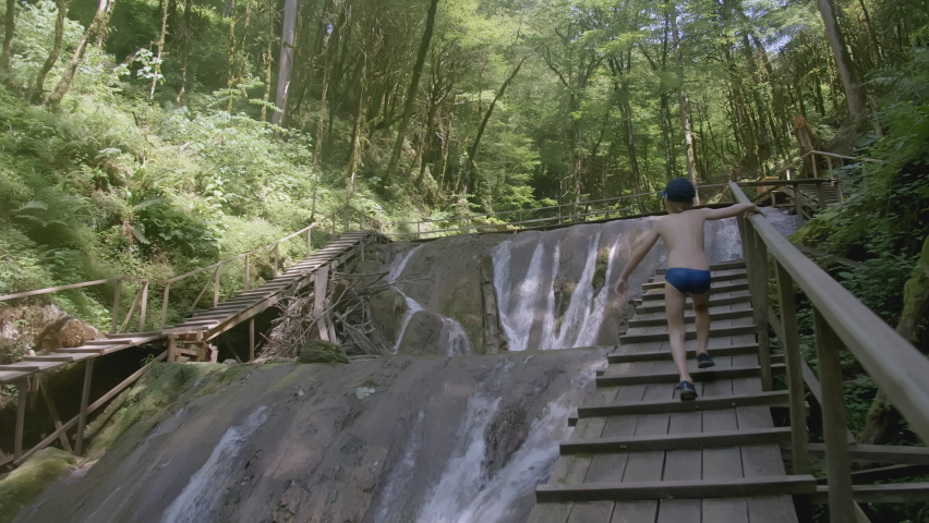 The boy runs up the wooden stairs against the backdrop of a waterfall. CREATIVE. Wooden stairs and ceilings in front of the waterfall. In the tropical vegetation is a lake with a waterfall. The guy