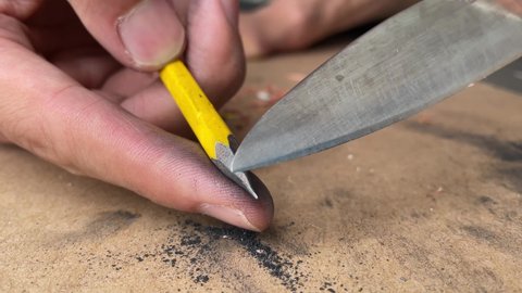 Male hand artist sharpening pencil with a sharp silver blade knife close up shot, educational and carpentry concept with cardboard box background - day