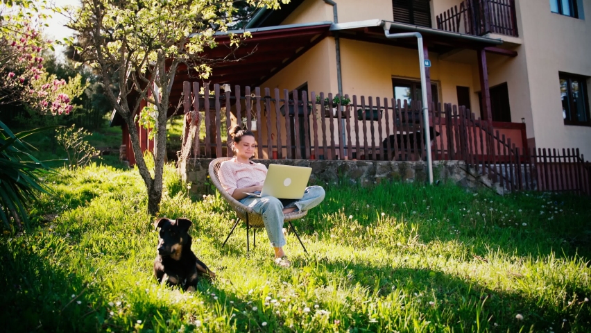 A young adult woman working on her laptop while sitting in the garden of her village house and her dog is lying on the lawn next to her.