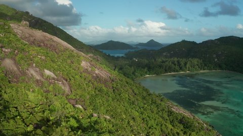 Aerial dolly slowly descending over the slope of hilly granite mountain with smooth boulders, covered green forests on the coast of Ternay bay with a beach in sunny weather with partly cloudy, Mahe