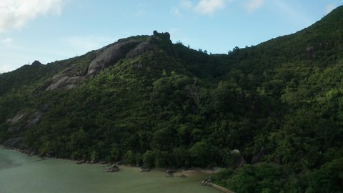 Aerial crane over Baie Ternay beach with green muddy water toward to the top of a granite rock with two huge boulders in the shadow of the clouds against the blue sky, Mahe, Seychelles