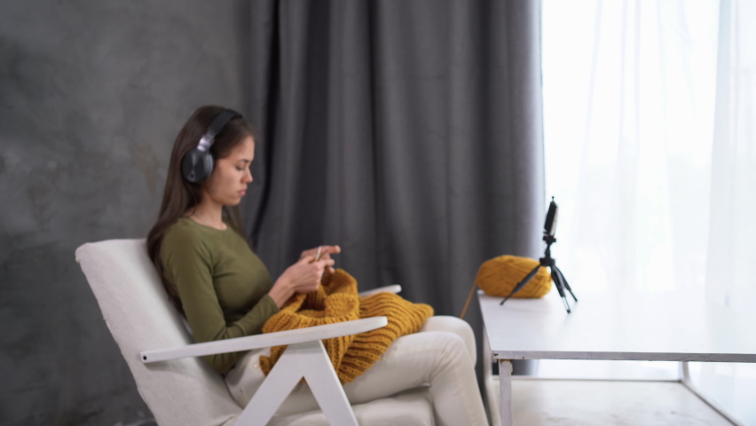 A modern woman in headphones is engaged in knitting at home thanks to sitting in a chair and watching a movie or series online using a smartphone.