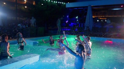 Friends have night pool party in a private villa swimming pool. Cheerful young people in swimwear splashing water, dancing and partying in luxury resort. Happy men and women hanging out. Slow motion.