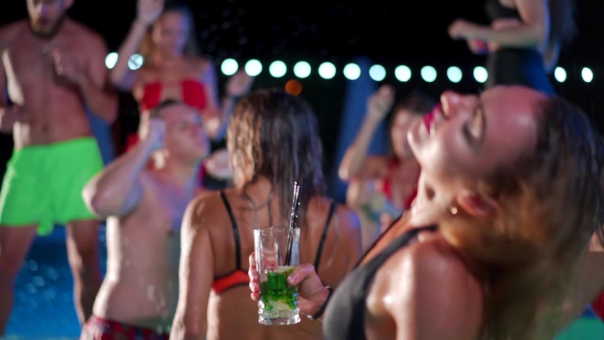 Hot woman in bikini drinking cocktail and hanging out with friends at night pool party. People having fun dancing with drinks and splashing water in luxury private villa swimming pool. Slow motion. Royalty-Free Stock Footage #1089810431