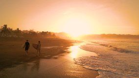 AERIAL SILHOUETTE: Two surfers walking down the beach carrying surfboards. Surf couple chatting in golden light before morning surf session. Gorgeous summer scenery at Playa Venao in Panama.