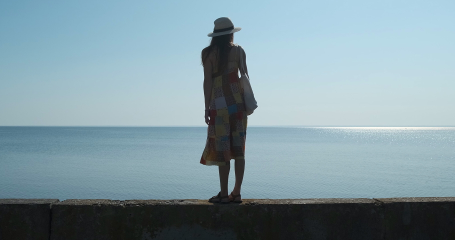 young woman in a hat stands on a concrete pier and looks at the calm sea in the distance. Daytime, calm, bright sun at its zenith, cloudless sky.