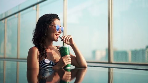 asian american woman in fashion swimsuit drinking non-alcoholic orange cocktail from straw, relaxation sitting in pool water, smiling at camera. Ethnic Girl enjoying vacation at luxury hotel in heat