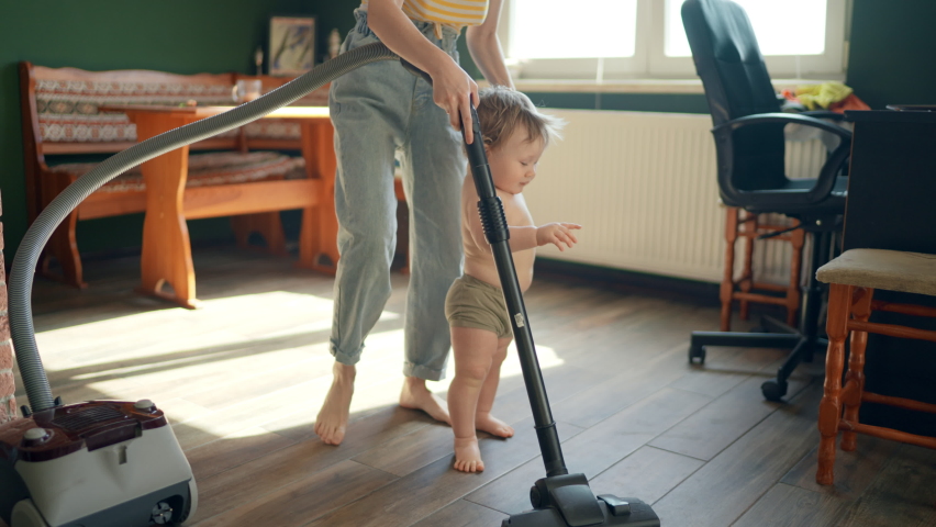 Mom and baby make vacuum cleaner cleaning in living room as mother and child play game indoors. Little feet walking on floor, Toddler learning to walk at home first steps. Smiling caring mother and