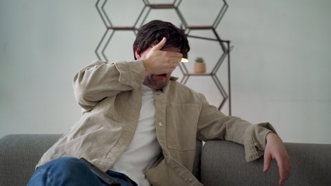 A man in the room holding a remote control while watching TV, pressing one palm to his face as if he is seeing a scary movie. 