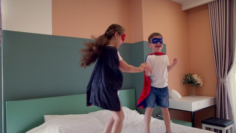 Superheroes, brother and sister, play at home imagining they are superheroes. Girl and boy superheroes, are jumping in room on bed,in children's room, two children in red and blue Superman costume,