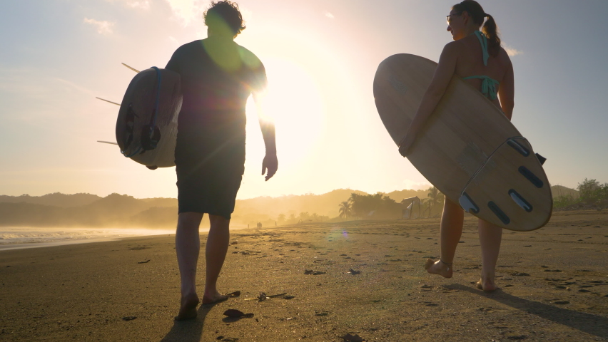 SLOW MOTION, LOW ANGLE VIEW: Couple of surfers walking on a sandy beach with surfboards. Two friends going surfing at sunset on Playa Venao surf spot. Beach lifestyle shot in golden light. Royalty-Free Stock Footage #1089823757