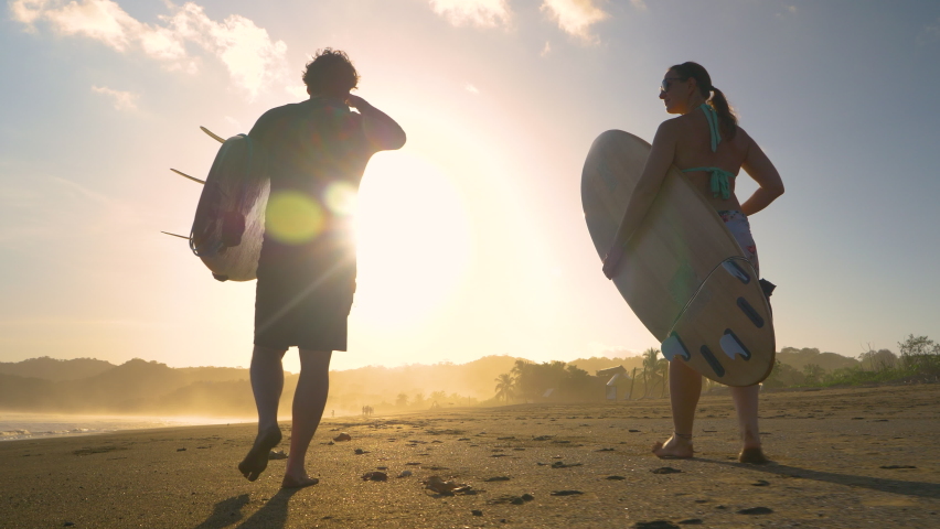SLOW MOTION, LOW ANGLE VIEW: Couple of surfers walking on a sandy beach with surfboards. Two friends going surfing at sunset on Playa Venao surf spot. Beach lifestyle shot in golden light. | Shutterstock HD Video #1089823757