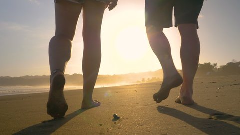 SLOW MOTION, LOW ANGLE VIEW: Following couple walking barefoot on sandy beach and leaving footprints. Two pairs of bare feet strolling along tropical seashore. Beach activity on summer holidays.