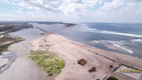 Esposende, Portugal, April 10, 2022: DRONE AERIAL FOOTAGE - The two sides of Restinga de Ofir. Esposende fishing harbor maintenance dredging. Dumper vehicles and rotating excavator parked on the sand.