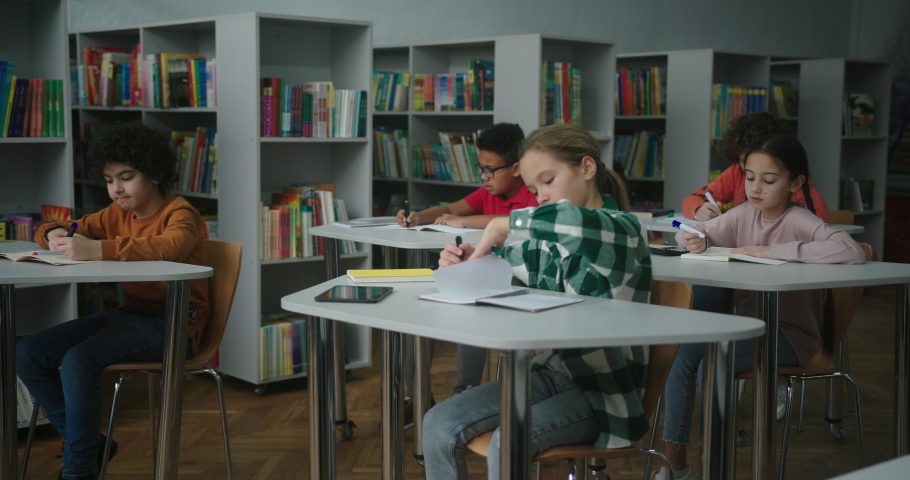 Schoolkids in protective masks perform task writing in exercise books at desks in library during pandemic. International children study at school slow motion Royalty-Free Stock Footage #1089826019