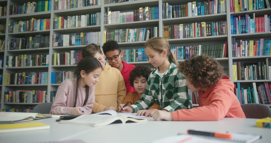 Multinational kids surround boy with kinky hair sitting at round table with stationery in library. Schoolchildren at extracurricular lesson slow motion Royalty-Free Stock Footage #1089826083