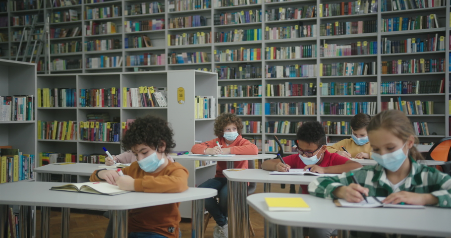 Schoolkids in protective masks perform task writing in exercise books at desks in library during pandemic. International children study at school slow motion Royalty-Free Stock Footage #1089826149