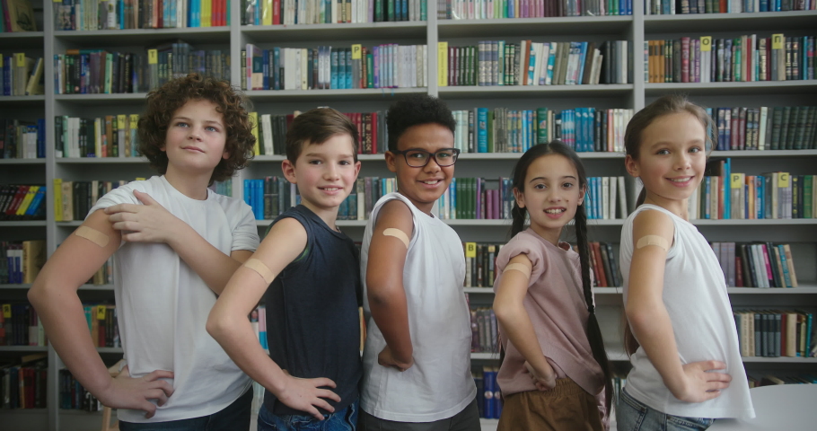 Happy international children make thumbs-up gesture after covid vaccination. Schoolkids show plasters on arms standing near books in library slow motion Royalty-Free Stock Footage #1089826173