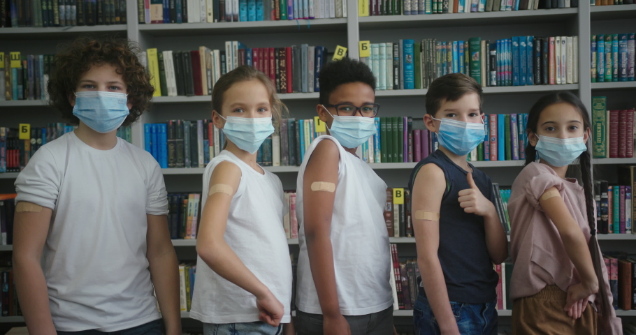 Multinational kids make thumbs-up gesture showing plasters on arms against bookshelves. Children in masks after vaccination in library slow motion Royalty-Free Stock Footage #1089826179