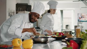 African american cook pouring shredded parmesan cheese on top of restaurant dish in frying pan. Male chef in uniform cooking professional gourmet meal with grated cheddar and ingredients in kitchen. - Powered by Shutterstock - Get 15% off with code: PIKWIZARD15
