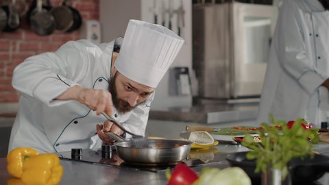 Male chef pouring shredded cheese on cooked food in frying pan, using grater and kitchen utensils to cook gourmet dish. Professional cook in white uniform adding grated parmesan on culinary meal.
