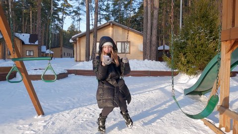 Teenage girl swinging on swing on playground and looking at mobile phone. Winter snowy cottages and pine trees background. Girl make selfie