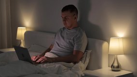 young man sitting on his bed and using laptop for video call in dark bedroom