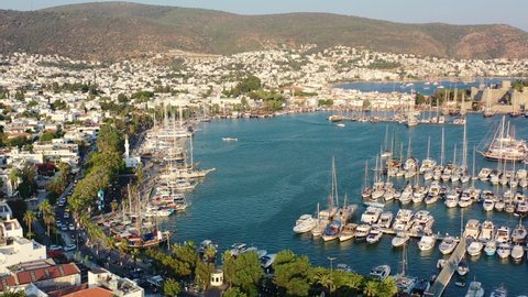 aerial drone flying across the Bodrum Marina with large sailboats docked in the Aegean Sea of Mugla Turkey as the sun sets over the hills and white villas on a summer afternoon