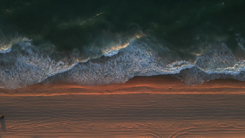 A stationary birdseye view shot of the shoreline with strong waves of water during the golden hour. The shoreline refers to the line where a body of water and the shore meet. The sand became tangerine
