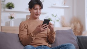 Positive asian guy using smartphone for online communication, scrolling news feed, messaging in social media, Man holds phone and smiling enjoy new mobile application on couch in living room at home.