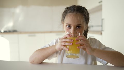 Little cute girl drinks fresh fruit juice. Happy child drinking orange drink. Healthy breakfast for a child. Girl drinks orange juice at the table in the kitchen. Healthy drink for a healthy lifestyle