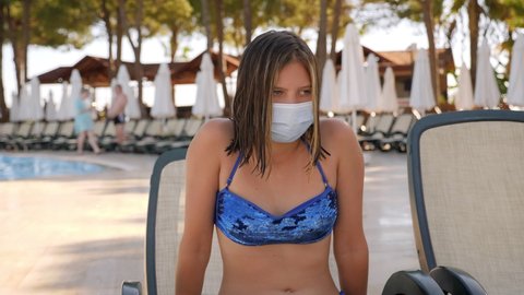 A teenage girl in a protective mask is relaxing by the pool on a chaise longue.