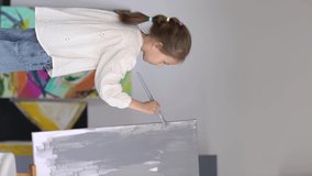 Young girl in white shirt and jeans painting big grey picture stands on the easel at home. Copy space. Vertical video. Art concept.