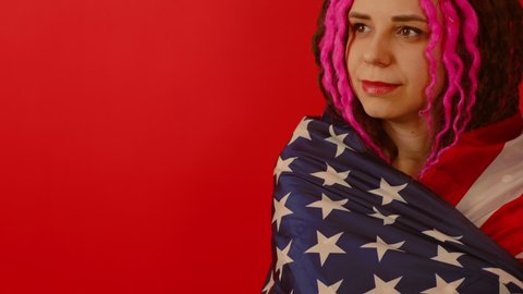 Young woman wrapped in American flag on red background with space for your text. Happy female smiling and looking away, posing in studio. Concept of Flag Day, national holiday.