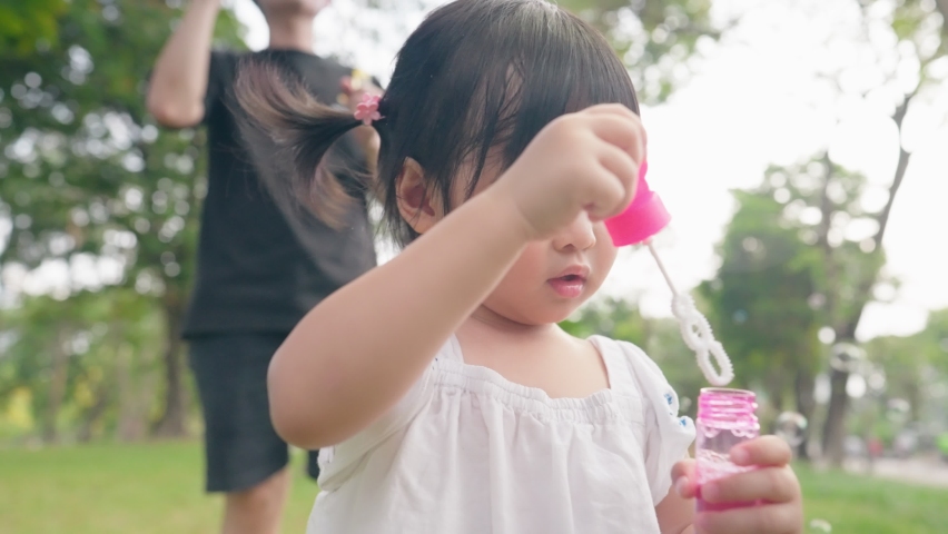 Asian girl blowing soap bubbles having fun playing with parents outside, Asian child innocent and curiosity, little preschool kid, family enjoy happy time in the park under trees shades Royalty-Free Stock Footage #1089845965