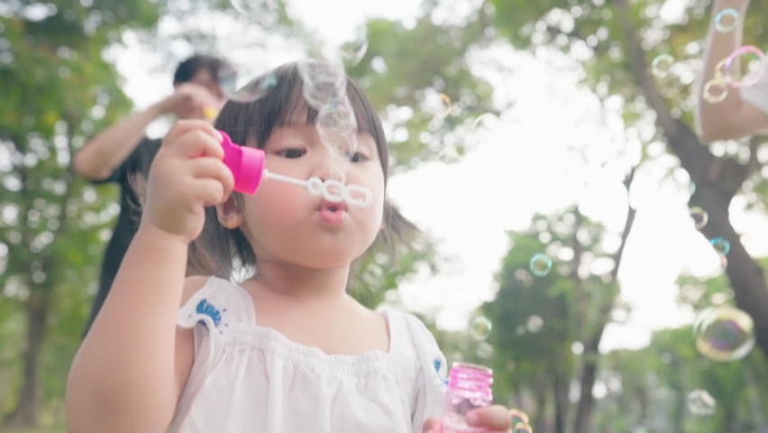 Asian girl blowing soap bubbles having fun playing with parents outside, Asian child innocent and curiosity, little preschool kid, family enjoy happy time in the park under trees shades Royalty-Free Stock Footage #1089845965