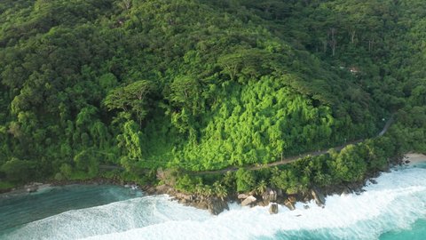 Aerial dolly over green blanket of invasive Kudzu vines, massive jungle trees and Indian ocean shore with granite boulders and winding road in the light of the setting sun, northwest coast of Mahe