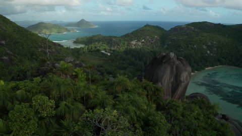 Aerial slider over mountain top near large granite boulder, palm trees and a bare tree overlooking the bay and beach Baie Ternay, and Therese island in cloudy weather with sunny gaps, Mahe, Seychelles