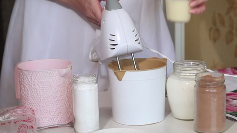 A woman mixes the ingredients to make a cream with a mixer. Adds condensed milk to container. The ingredients are on the table. Preparing eclairs. Medium plan.