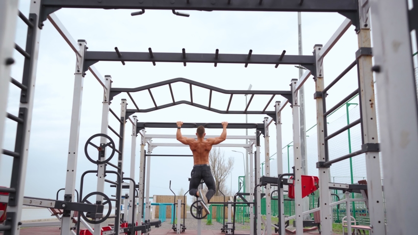 Athletic Man Pumps Up Back Muscles Doing Pull-Ups in an Open Sports Ground with a Bare Torso. Muscular Handsome Guy is Engaged in Workout on Horizontal Bar. Fitness and Bodybuilding Concept Outdoors. Royalty-Free Stock Footage #1089848485