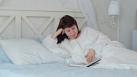Middle shot of a woman in white nightgown lies on bed in bedroom and reads a book in the morning with daylight, 4k video