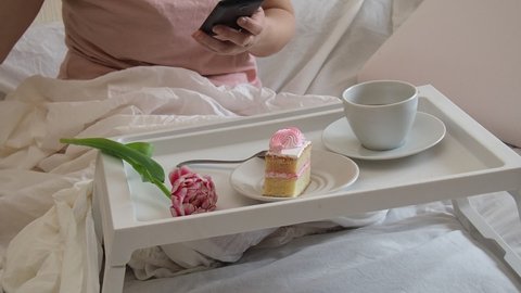 Cheerful tender female in white sleepwear sitting with cup of coffee on bed and having breakfast while enjoying morning in bedroom at home. watches news on the phone, social networks, eats cake